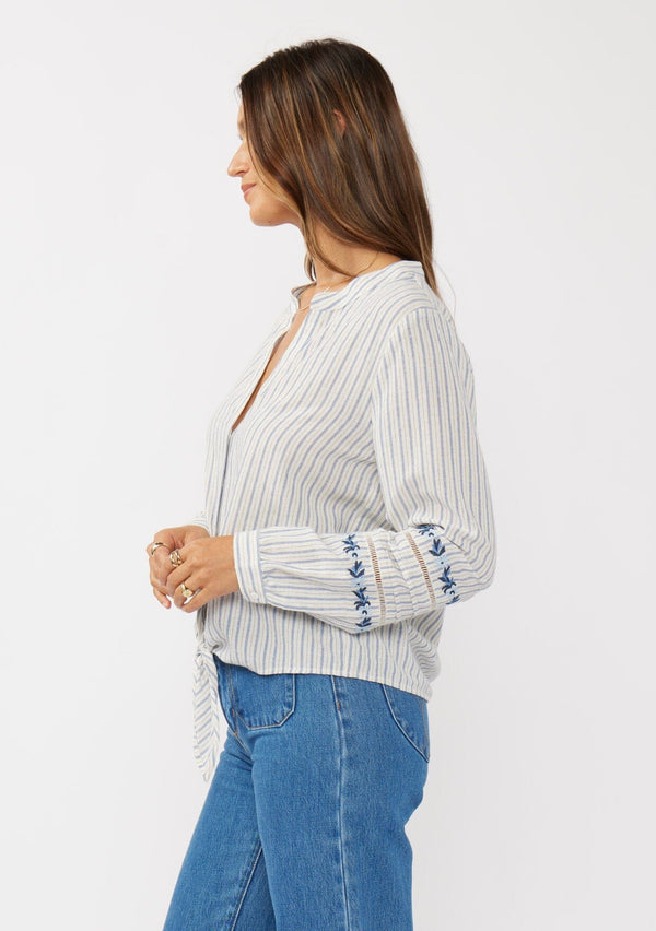 [Color: Navy/Natural] A side facing image of a brunette model wearing a blue yarn dye stripe blouse with a floral embroidery,  tie front, long sleeves with button cuffs, and a split v-neckline.