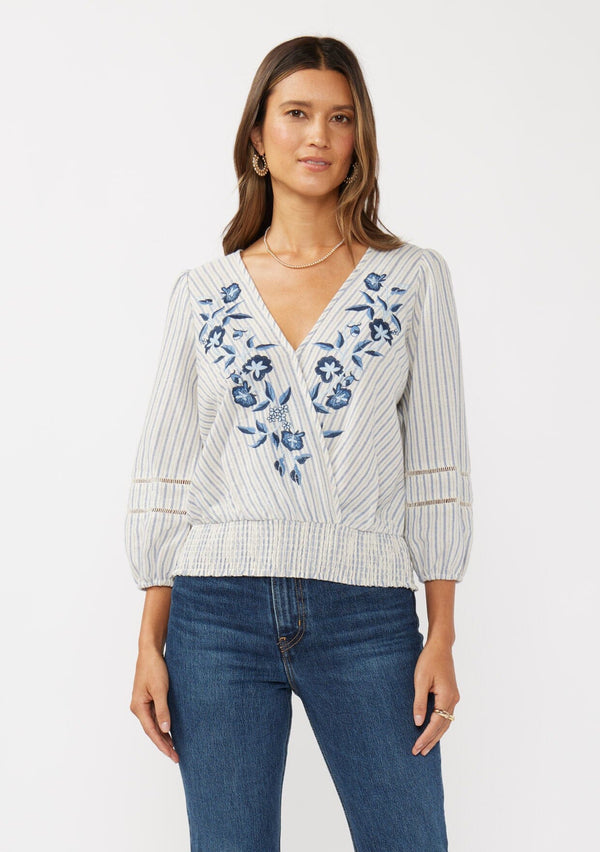 [Color: Navy/Natural] A front facing image of a brunette model wearing a blue yarn dye stripe top with a floral embroidery, surplice v-neckline, smocked hem, and 3/4 length sleeves. 