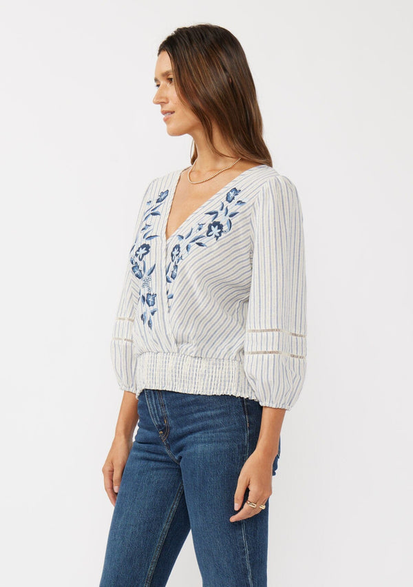 [Color: Navy/Natural] A side facing image of a brunette model wearing a blue yarn dye stripe top with a floral embroidery, surplice v-neckline, smocked hem, and 3/4 length sleeves. 