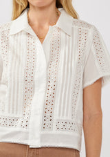 [Color: White] A detail image of a blonde model wearing a white floral eyelet shirt in a cropped length. This buttoned shirt features a button front, collared neckline, pintuck details, and picot trim. The perfect resort top for layering. 