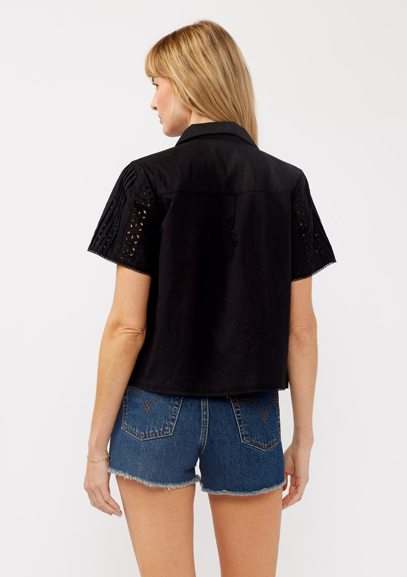 [Color: Black] A back facing image of a blonde model wearing a black floral eyelet shirt in a cropped length. This unbuttoned shirt features a button front, collared neckline, pintuck details, and picot trim. The perfect resort top for layering. 