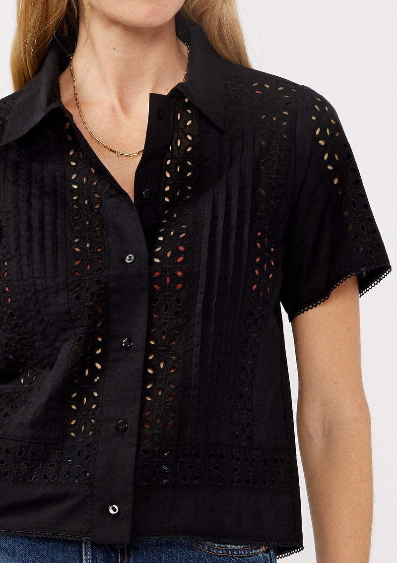 [Color: Black] A detail image of a blonde model wearing a black floral eyelet shirt in a cropped length. This unbuttoned shirt features a button front, collared neckline, pintuck details, and picot trim. The perfect resort top for layering. 