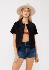 [Color: Black] A front facing image of a blonde model wearing a black floral eyelet shirt in a cropped length. This unbuttoned shirt features a button front, collared neckline, pintuck details, and picot trim. The perfect resort top for layering. 