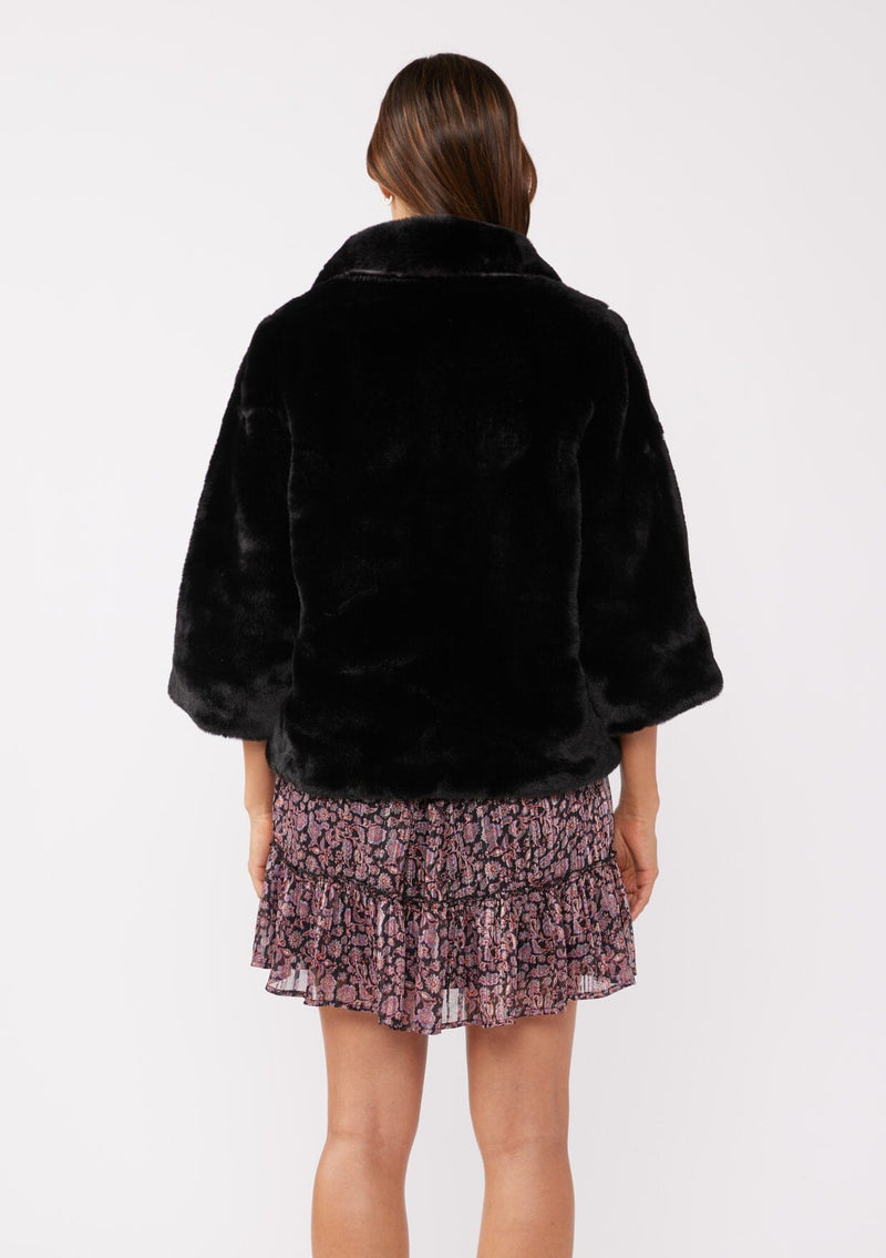 [Color: Black] A back facing image of a brunette model wearing a black faux fur coat with ¾ length sleeves and a collar. A cozy holiday statement piece with a relaxed fit, perfect for layering over a dress. Versatile and timeless.