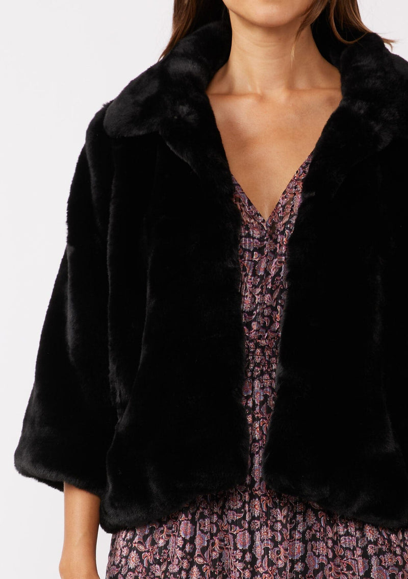 [Color: Black] A brunette model wearing a black faux fur coat with ¾ length sleeves and a collar. A cozy holiday statement piece with a relaxed fit, perfect for layering over a dress. Versatile and timeless.