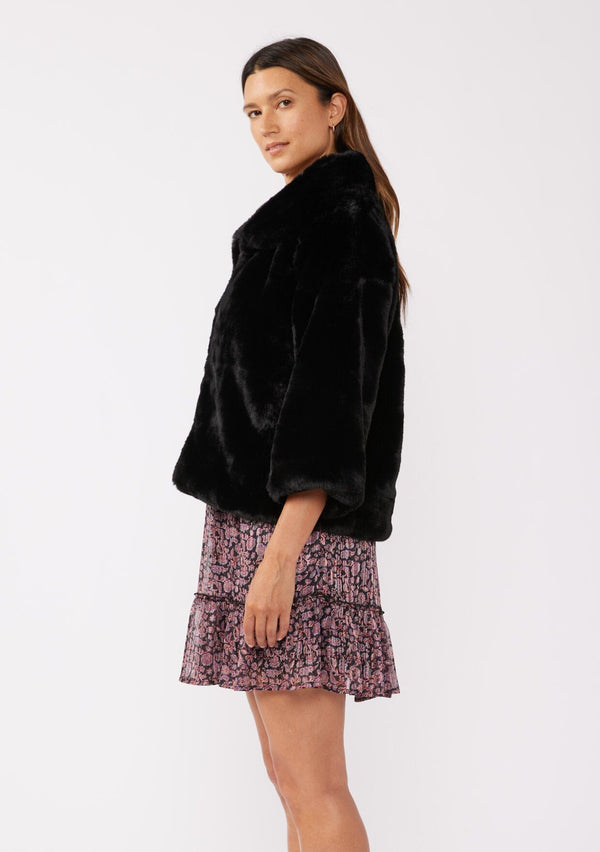 [Color: Black] A side facing image of a brunette model wearing a black faux fur coat with ¾ length sleeves and a collar. A cozy holiday statement piece with a relaxed fit, perfect for layering over a dress. Versatile and timeless.