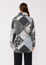 [Color: Natural/Blue] A back facing image of a brunette model wearing a blue and white patchwork print quilted jacket. With long sleeves, a shawl collar, side pockets, and a tie waist belt. Styled with a turtleneck and trousers for a formal yet casual look. A fall season jacket.  