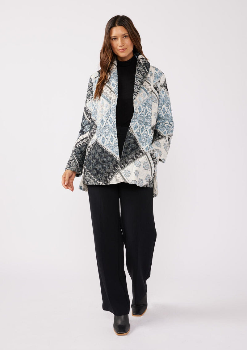 [Color: Natural/Blue] A front facing image of a brunette model wearing a blue and white patchwork print quilted jacket. With long sleeves, a shawl collar, side pockets, and a tie waist belt. Styled with a turtleneck and trousers for a formal yet casual look. A fall season jacket.  