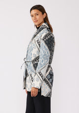 [Color: Natural/Blue] A side facing image of a brunette model wearing a blue and white patchwork print quilted jacket. With long sleeves, a shawl collar, side pockets, and a tie waist belt. Styled with a turtleneck and trousers for a formal yet casual look. A fall season jacket.  