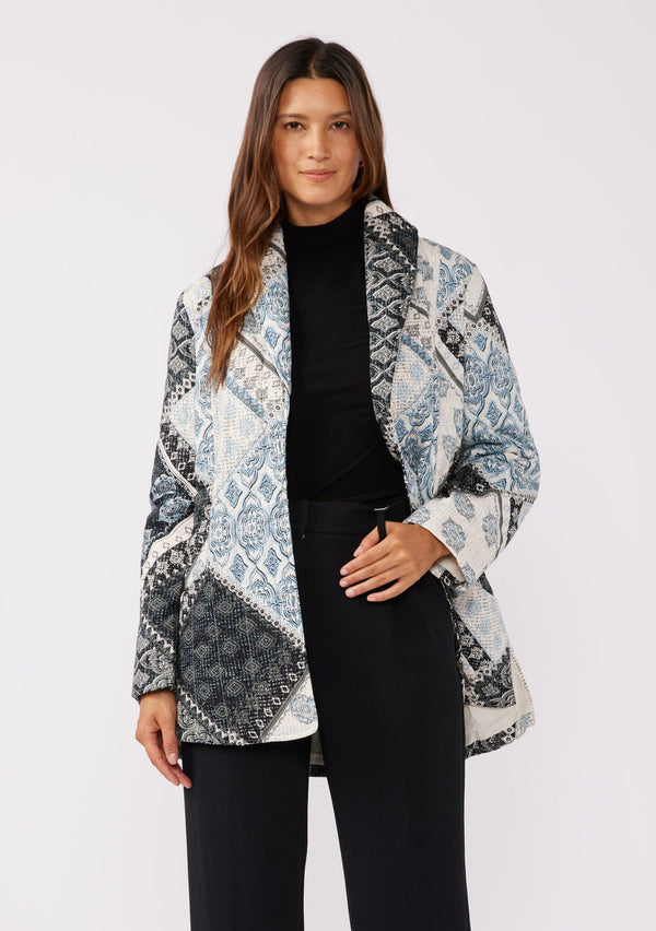 [Color: Natural/Blue] A front facing image of a brunette model wearing a blue and white patchwork print quilted jacket. With long sleeves, a shawl collar, side pockets, and a tie waist belt. Styled with a turtleneck and trousers for a formal yet casual look. A fall season jacket.  