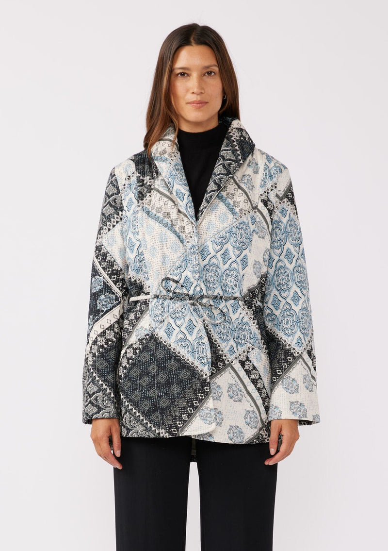 [Color: Natural/Blue] A front facing image of a brunette model wearing a blue and white patchwork print quilted jacket. With long sleeves, a shawl collar, side pockets, and a tie waist belt. Styled with a turtleneck and trousers for a formal yet casual look. A fall season jacket.  