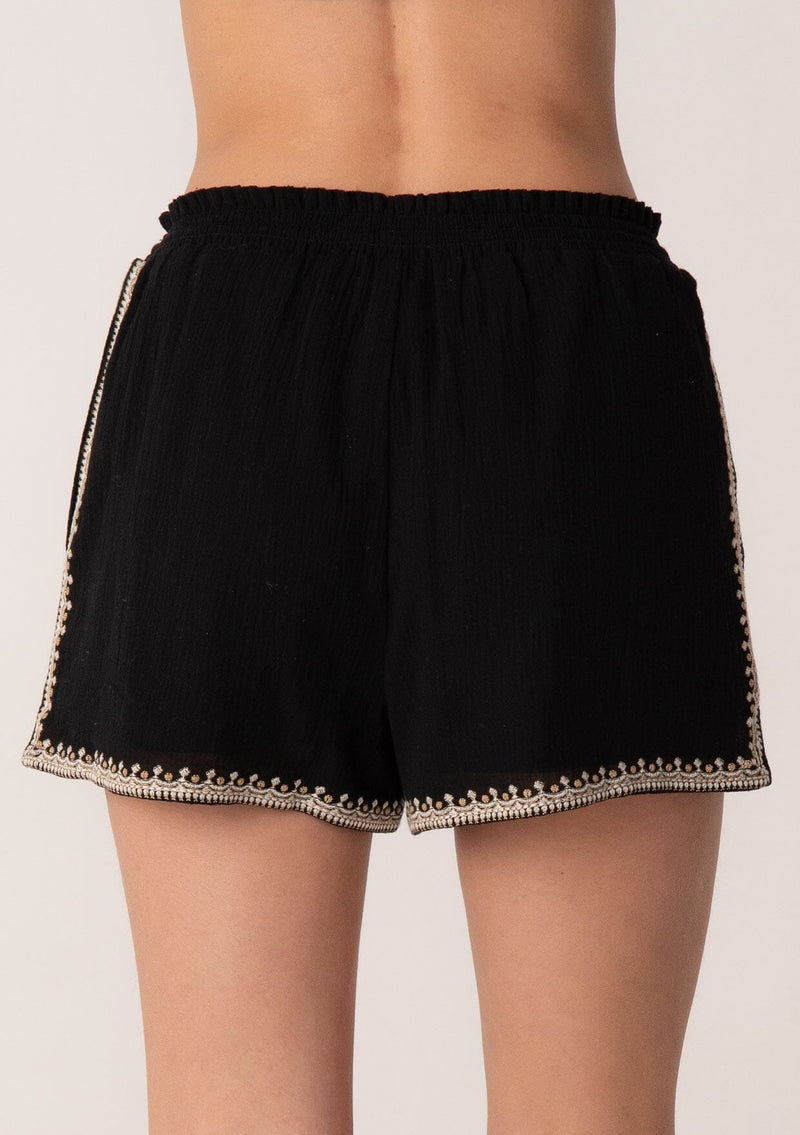 [Color: Black/Natural] A close up back facing image of a blonde model wearing a black bohemian cotton short with embroidered detail. With a smocked elastic waist, a drawstring tie waist, and side pockets.