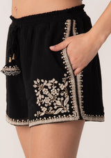 [Color: Black/Natural] A close up side facing image of a blonde model wearing a black bohemian cotton short with embroidered detail. With a smocked elastic waist, a drawstring tie waist, and side pockets.
