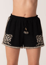 [Color: Black/Natural] A close up front facing image of a blonde model wearing a black bohemian cotton short with embroidered detail. With a smocked elastic waist, a drawstring tie waist, and side pockets.