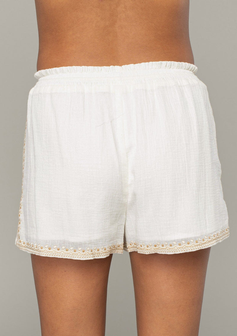 [Color: Natural/Taupe] A close up back facing image of a brunette model wearing an off white bohemian cotton short with embroidered detail. With a smocked elastic waist, a drawstring tie waist, and side pockets.