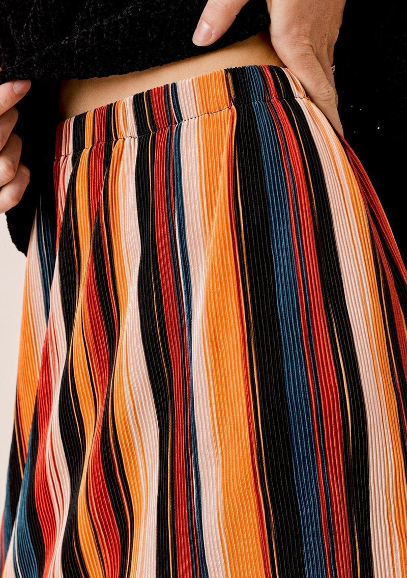 [Color: Black/Spice/Teal:] A detailed image of a blonde wearing a colorful pleated midi skirt with an elastic waistband and front slit. Perfectly versatile, this vertical stripe skirt is styled with a cozy sweater for a laid back look, perfect for the Fall season. 