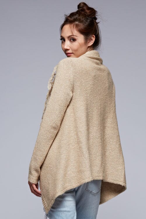 [Color: Oatmeal] A back facing image of a brunette model wearing a cozy one-sided fringe sweater cardigan with a versatile wrap front. This asymmetrical sweater features two side buttons for easy draping and securing the wrap silhouette. 