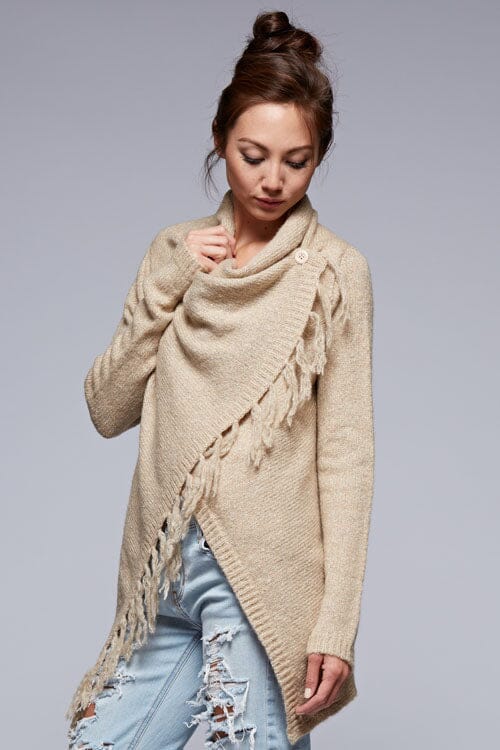 [Color: Oatmeal] A side facing image of a brunette model wearing a cozy one-sided fringe sweater cardigan with a versatile wrap front. This asymmetrical sweater features two side buttons for easy draping and securing the wrap silhouette. 