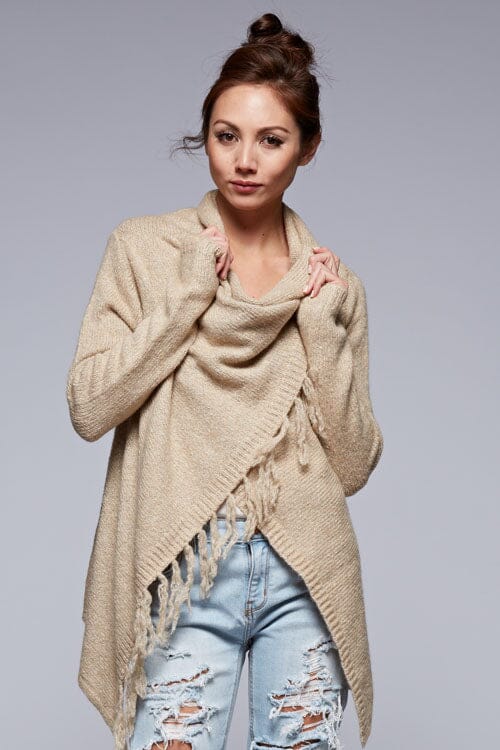 [Color: Oatmeal] A front facing image of a brunette model wearing a cozy one-sided fringe sweater cardigan with a versatile wrap front. This asymmetrical sweater features two side buttons for easy draping and securing the wrap silhouette. 