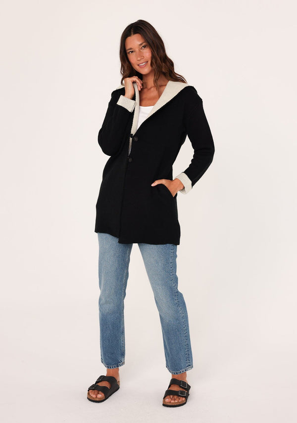 [Color: Black/Natural] A brunette model wearing a black sweater coat with a contrast interior, front button snap closure, side pockets, and a hoodie. Perfect for laid-back outings or lounging at home. 