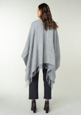 [Color: Heather Grey] A back facing image of a brunette model wearing a throw-on grey blanket poncho with an open design, long length, and fringe trim. Perfect for effortless cold weather styling.