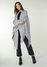 [Color: Heather Grey] A front facing image of a brunette model wearing a throw-on grey blanket poncho with an open design, long length, and fringe trim. Perfect for effortless cold weather styling.