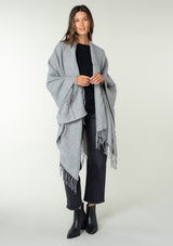 [Color: Heather Grey] A front facing image of a brunette model wearing a throw-on grey blanket poncho with an open design, long length, and fringe trim. Perfect for effortless cold weather styling.