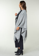 [Color: Heather Grey] A side facing image of a brunette model wearing a throw-on grey blanket poncho with an open design, long length, and fringe trim. Perfect for effortless cold weather styling.