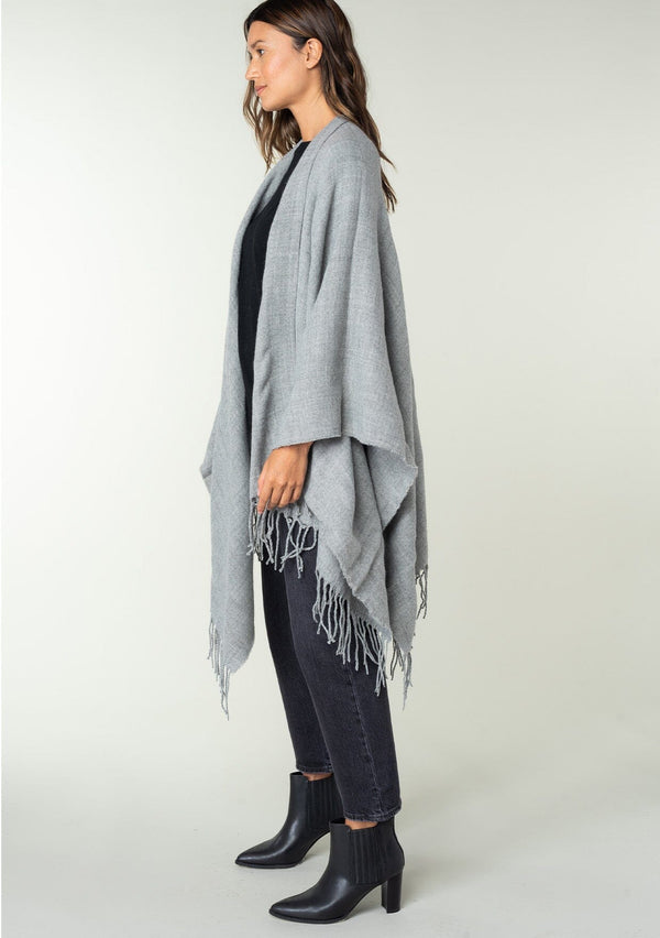 [Color: Heather Grey] A side facing image of a brunette model wearing a throw-on grey blanket poncho with an open design, long length, and fringe trim. Perfect for effortless cold weather styling.