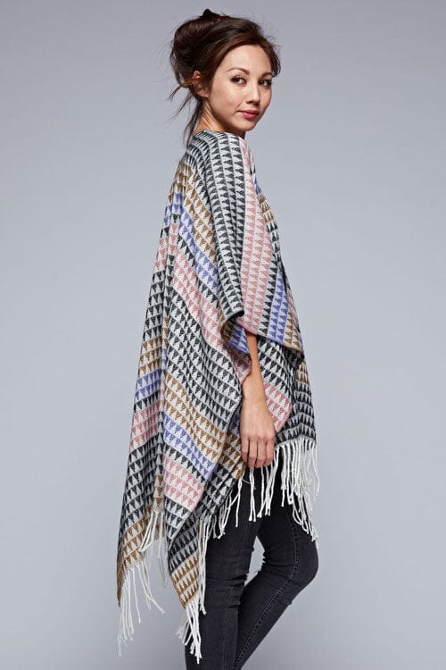 [Color: Black/Natural] A side facing image of a brunette model wearing a geometric stripe knit bohemian poncho with an open front and fringe trim. 