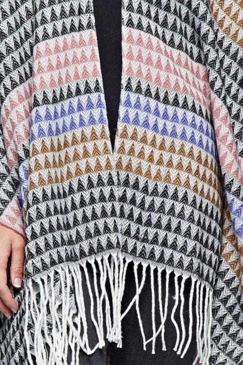 [Color: Black/Natural] A detail image of a brunette model wearing a geometric stripe knit bohemian poncho with an open front and fringe trim. 