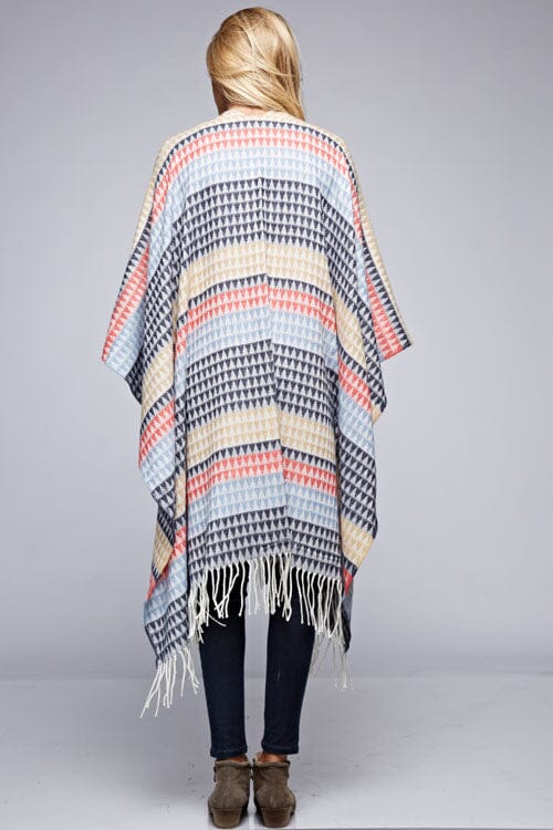 [Color: Navy/Vanilla] A back facing image of a blonde model wearing a geometric stripe knit bohemian poncho with an open front and fringe trim. 