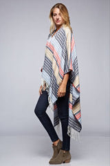 [Color: Navy/Vanilla] A side facing image of a blonde model wearing a geometric stripe knit bohemian poncho with an open front and fringe trim. 