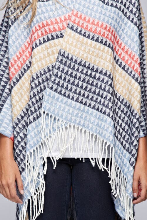 [Color: Navy/Vanilla] A detail image of a blonde model wearing a geometric stripe knit bohemian poncho with an open front and fringe trim. 