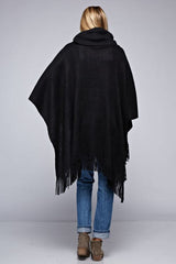 [Color: Black] A back facing image of a blonde model wearing a black ultra cozy poncho crafted in a soft acrylic. Featuring an exaggerated cowl neckline, asymmetrical hemline, and fringe trim details. 