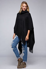 [Color: Black] A side facing image of a blonde model wearing a black ultra cozy poncho crafted in a soft acrylic. Featuring an exaggerated cowl neckline, asymmetrical hemline, and fringe trim details. 