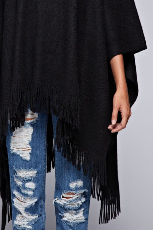 [Color: Black] A detail image of a blonde model wearing a black ultra cozy poncho crafted in a soft acrylic. Featuring an exaggerated cowl neckline, asymmetrical hemline, and fringe trim details. 