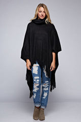 [Color: Black] A front facing image of a blonde model wearing a black ultra cozy poncho crafted in a soft acrylic. Featuring an exaggerated cowl neckline, asymmetrical hemline, and fringe trim details. 