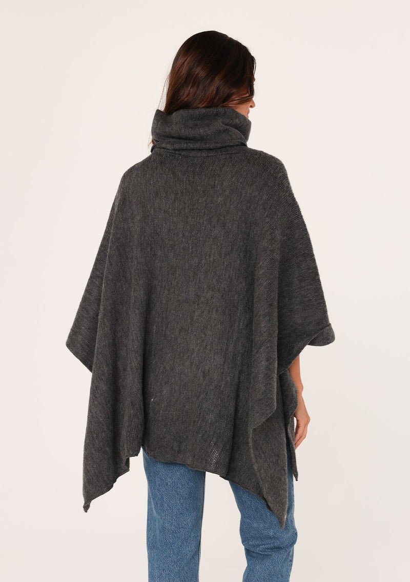 [Color: Dark Charcoal] A brunette model wearing a cozy acrylic knit grey poncho with a cowl neckline and asymmetrical hemline. Perfect for cozy winters and fall styling. 