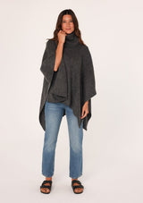 [Color: Dark Charcoal] A brunette model wearing a cozy acrylic knit grey poncho with a cowl neckline and asymmetrical hemline. Perfect for cozy winters and fall styling. 