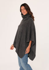 [Color: Dark Charcoal] A brunette model wearing a cozy acrylic knit grey poncho with a cowl neckline and asymmetrical hemline. Perfect for cozy winters and fall styling. 