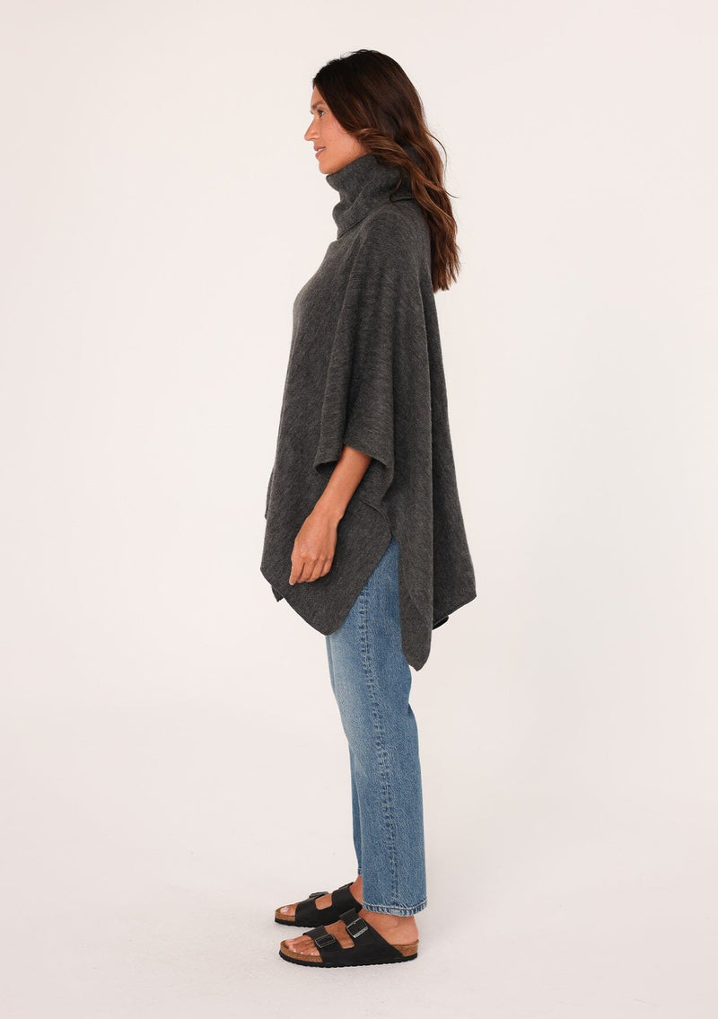 [Color: Dark Charcoal] A brunette model wearing a cozy acrylic knit grey poncho with a cowl neckline and asymmetrical hemline. Perfect for cozy winters and fall styling. 