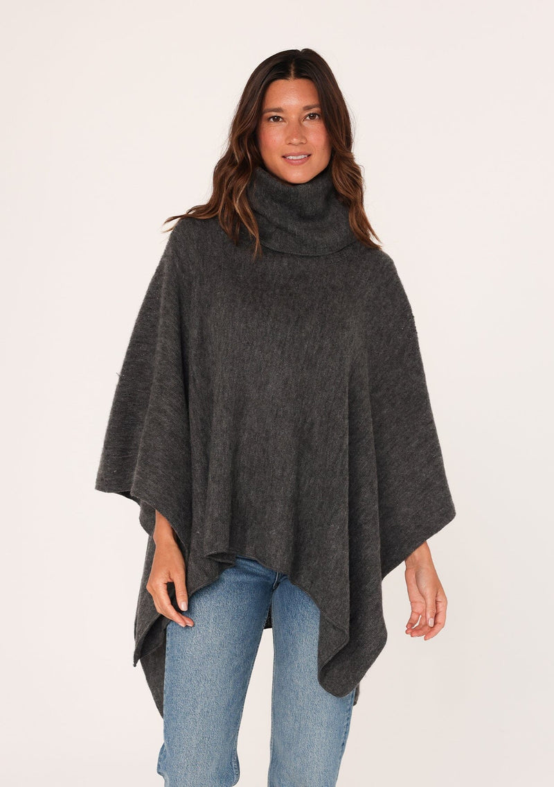 [Color: Dark Charcoal] A brunette model wearing a cozy acrylic knit grey poncho with a cowl neckline and asymmetrical hemline. Perfect for cozy winters and fall styling. 