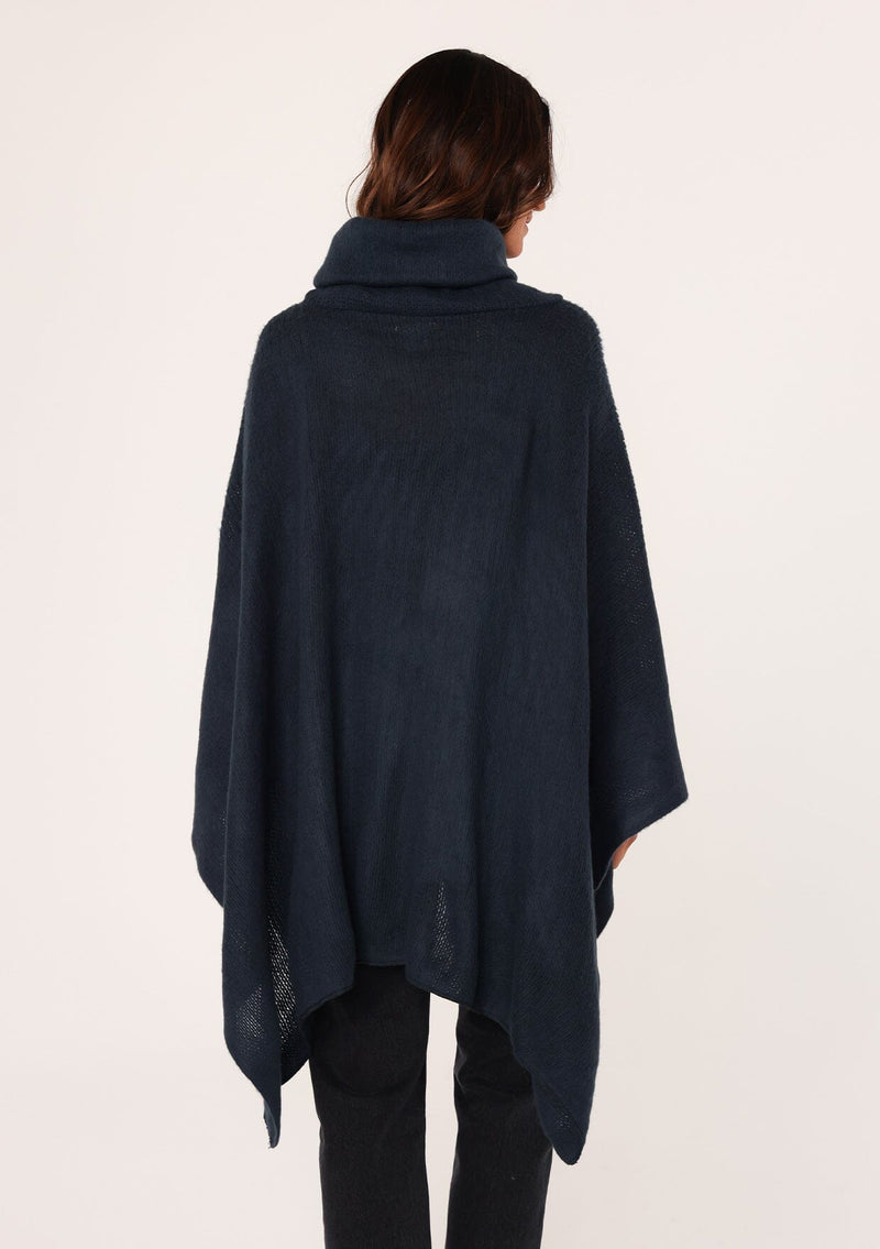 [Color: Navy] A brunette model wearing a cozy acrylic knit navy blue poncho with a cowl neckline and asymmetrical hemline. Perfect for cozy winters and fall styling. 