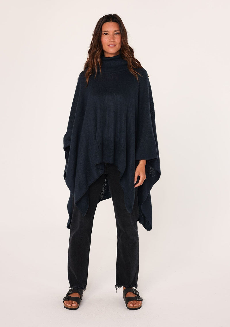 [Color: Navy] A brunette model wearing a cozy acrylic knit navy blue poncho with a cowl neckline and asymmetrical hemline. Perfect for cozy winters and fall styling. 