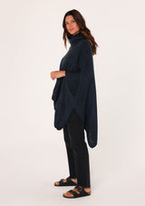 [Color: Navy] A brunette model wearing a cozy acrylic knit navy blue poncho with a cowl neckline and asymmetrical hemline. Perfect for cozy winters and fall styling. 