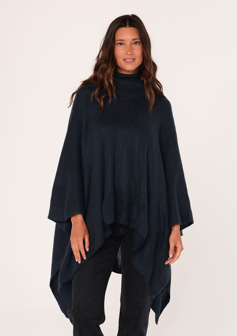 [Color: Navy] A brunette model wearing a cozy acrylic knit navy blue poncho with a cowl neckline and asymmetrical hemline. Perfect for cozy winters and fall styling. 