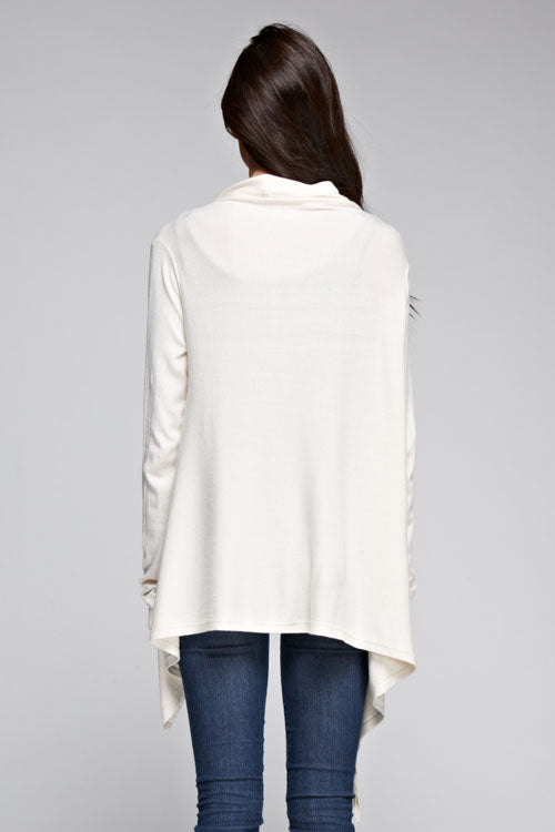 [Color: Vanilla] A brunette model wearing an off white asymmetrical wrap front cardigan with a suede fringe trim and long sleeves with thumb holes. Perfect for elevating any casual outfit. 