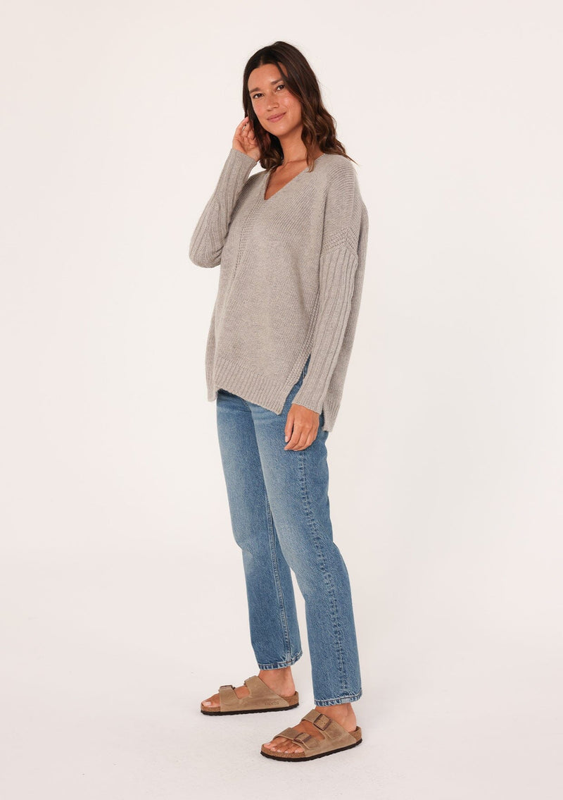 [Color: Heather Grey] A side facing image of a brunette model wearing a cozy grey pullover sweater with a v-neckline, ribbed long sleeves, dropped shoulders, and side vents. The perfect throw on for fall styling. 