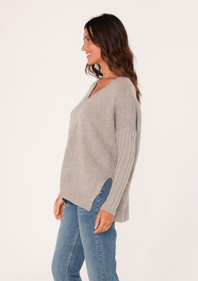 [Color: Heather Grey] A side facing image of a brunette model wearing a cozy grey pullover sweater with a v-neckline, ribbed long sleeves, dropped shoulders, and side vents. The perfect throw on for fall styling. 